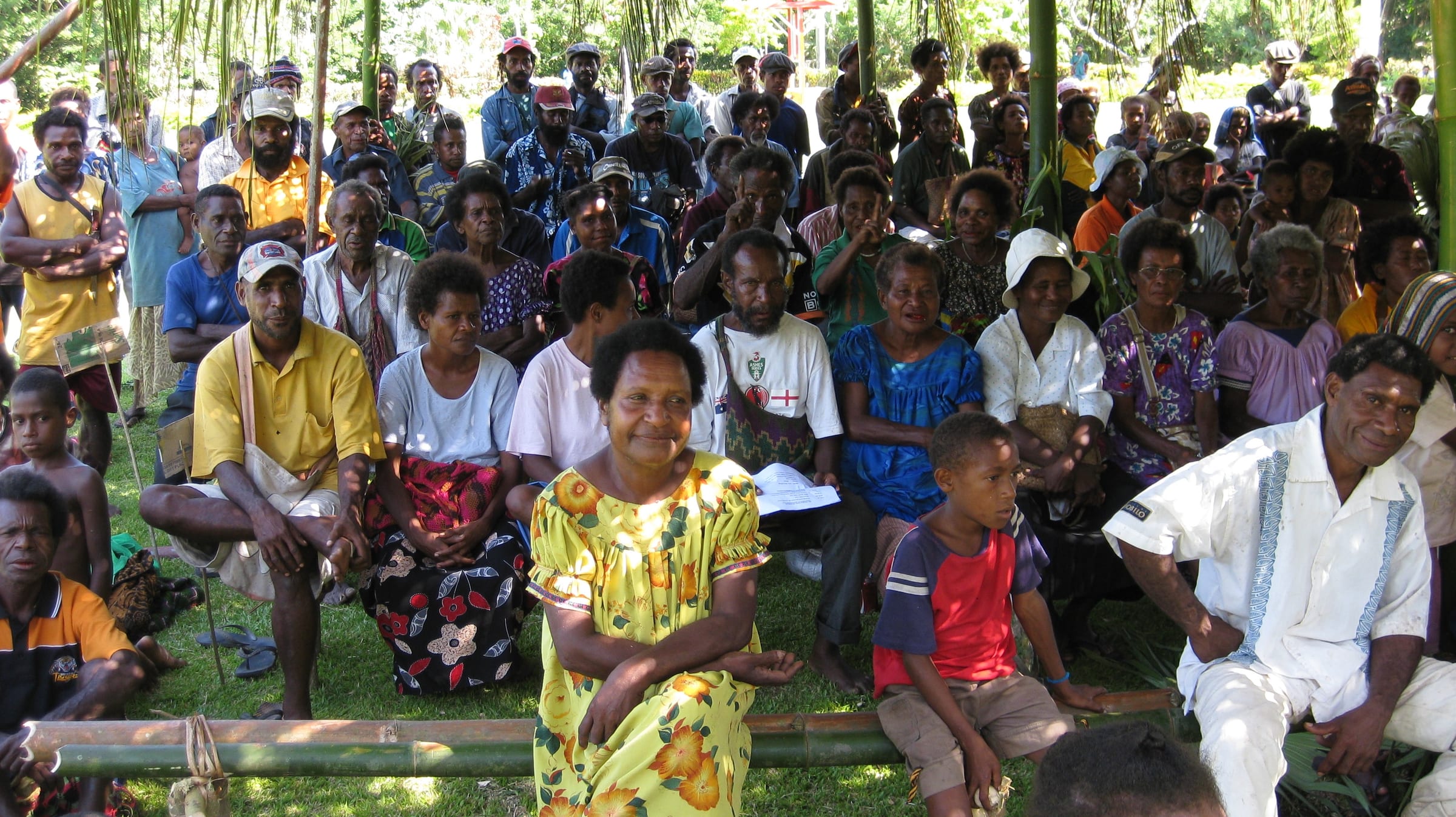 Villagers of all ages seated together under a bush-material shelter at a community meeting in Papua New Guinea